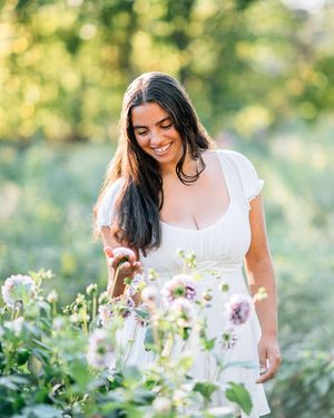 Person in white dress smiling while tending to purple wildflowers in a sunlit garden with bokeh background.