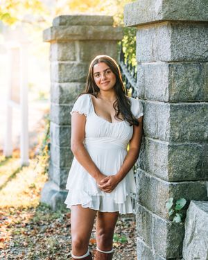 A white summer dress is worn against a stone pillar backdrop in warm sunlight with autumn leaves on the ground.
