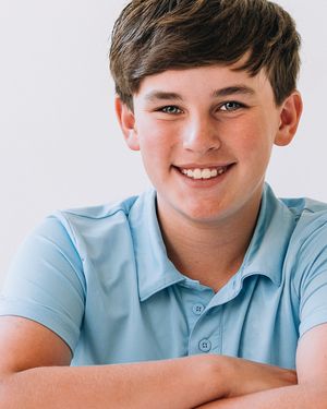 Smiling young boy with brown hair wearing a light blue polo shirt, arms crossed, against a white background.