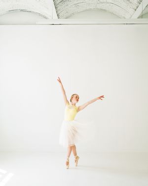 Ballet dancer in yellow top and white skirt gracefully poses en pointe against bright minimalist studio background.