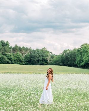 A woman in a white maxi dress stands in a blooming wildflower field with lush green trees under cloudy skies.