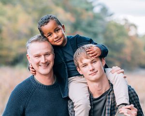 Three smiling males pose outdoors in autumn, with a child sitting on the shoulders of two others.