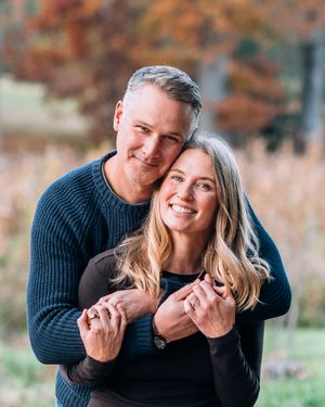 A smiling couple embraces outdoors in autumn, wearing matching navy sweaters with colorful fall foliage in the background.
