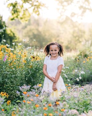 A smiling young girl in a white dress stands among colorful wildflowers in a sunlit garden.