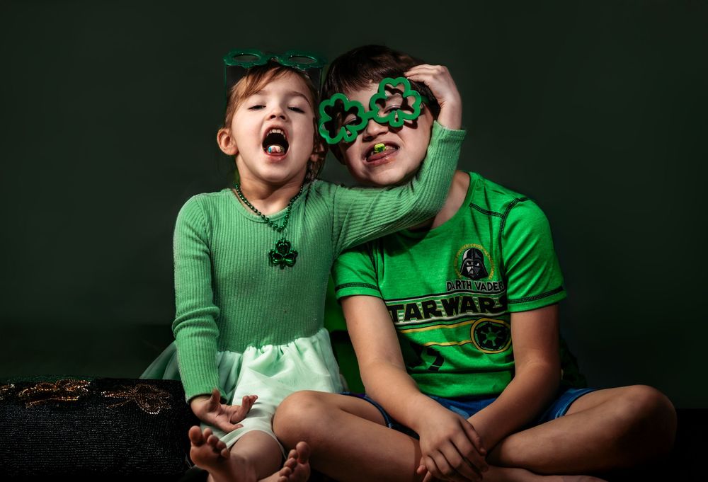 Two children in green shirts laugh while wearing St. Patrick's Day accessories against dark background.