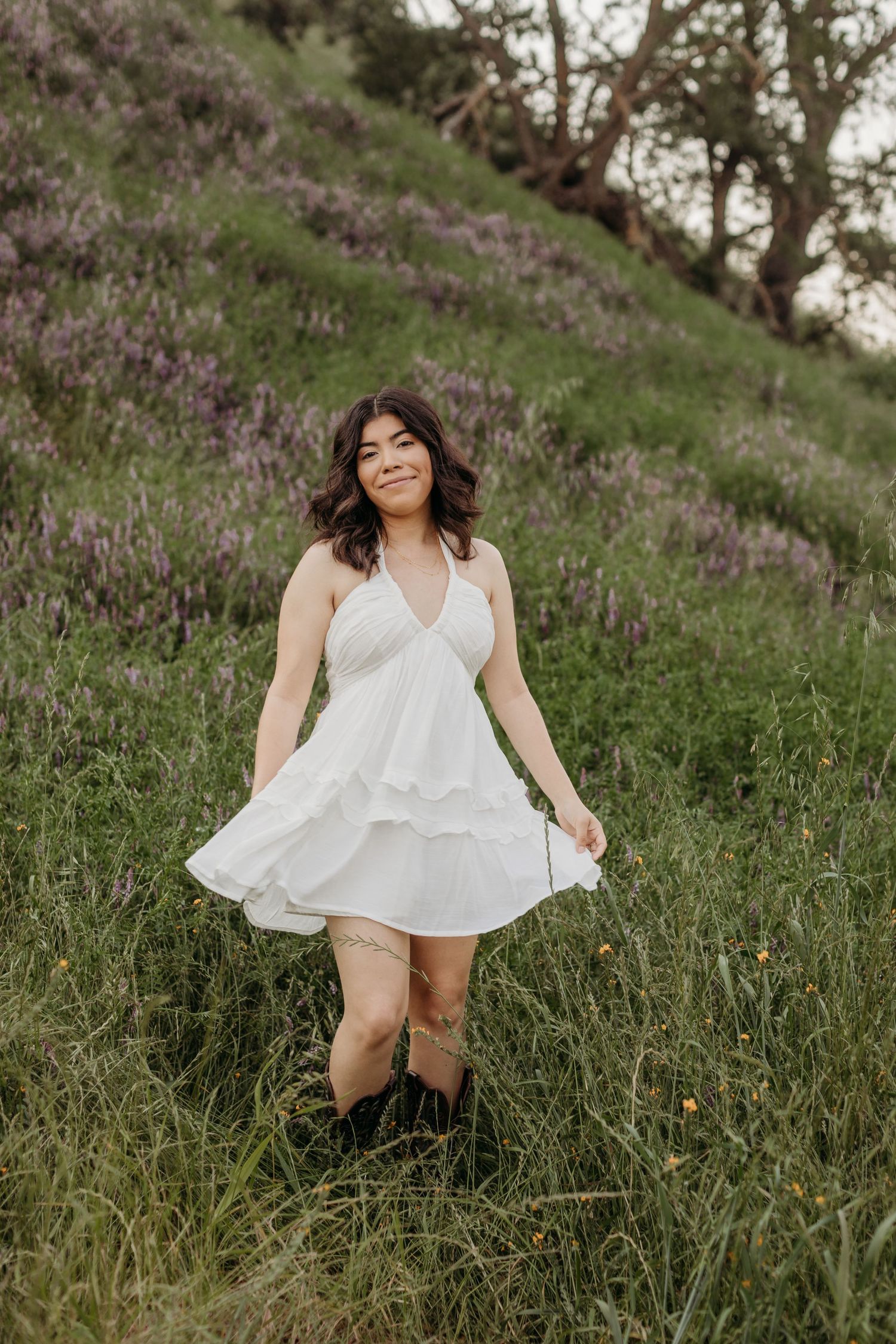 Woman in white sundress poses on hillside covered in purple wildflowers.
