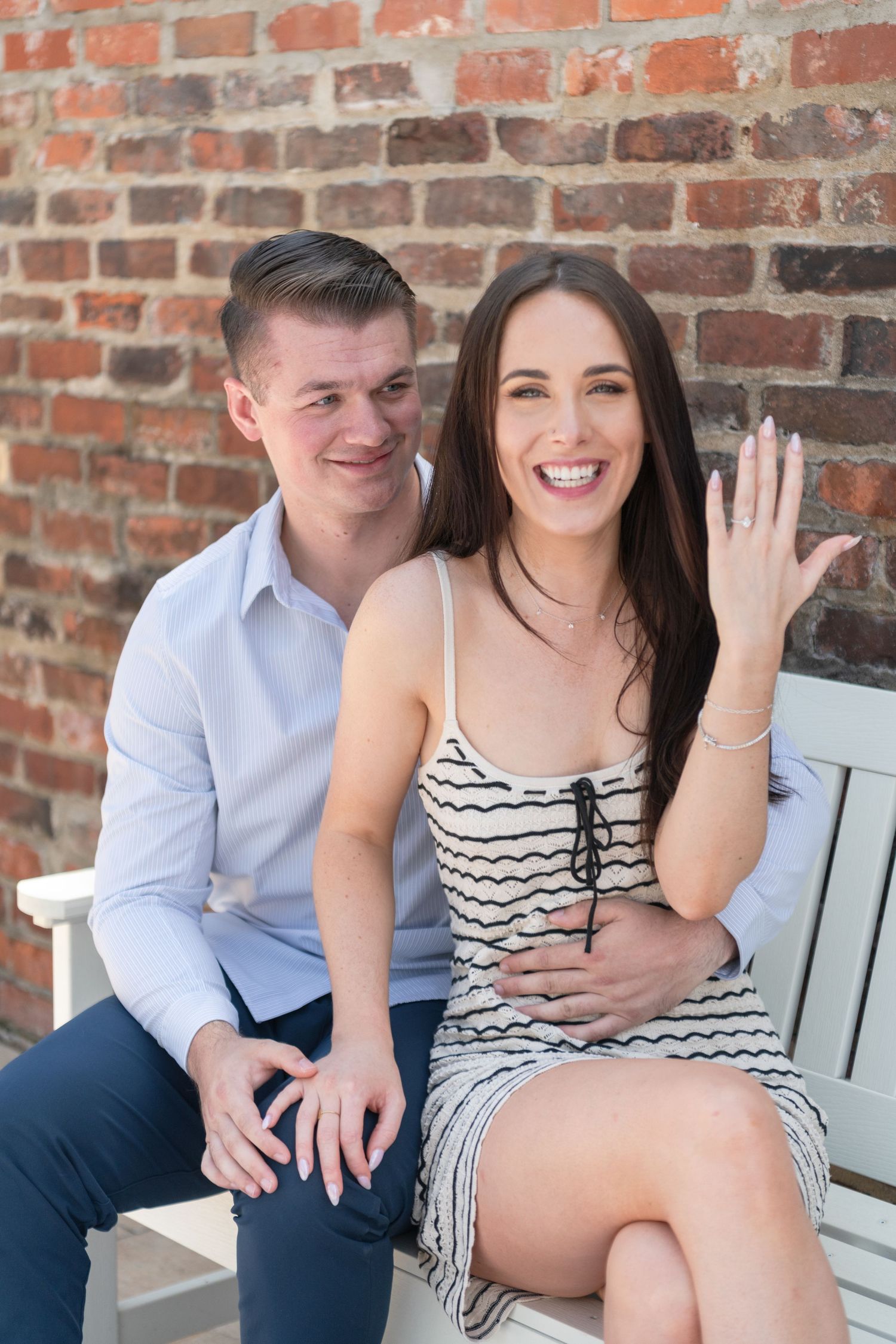 Couple sitting on white bench against brick wall showing off engagement ring.