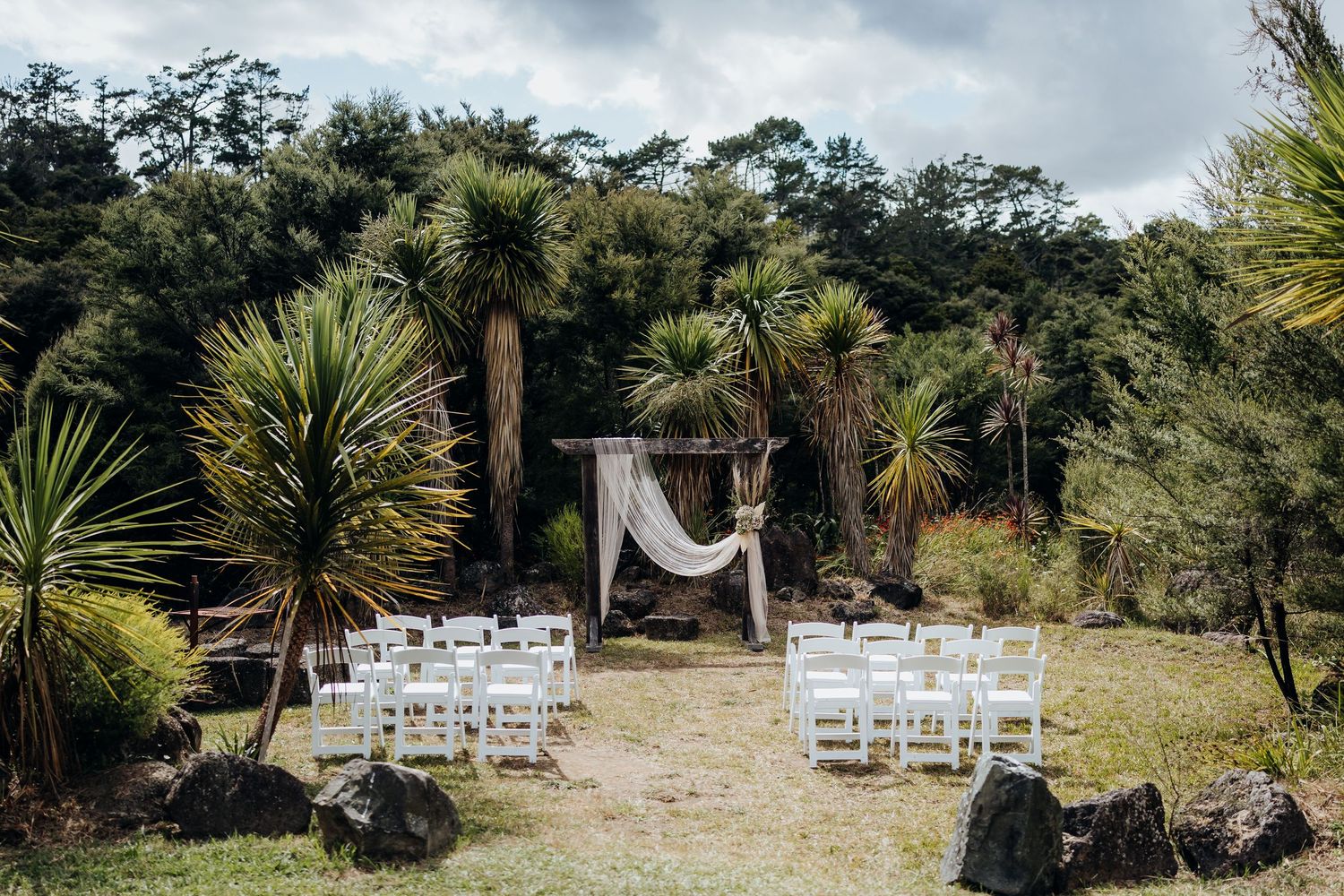 A tropical wedding ceremony setup with white chairs and fabric arch surrounded by palm trees and tropical foliage.