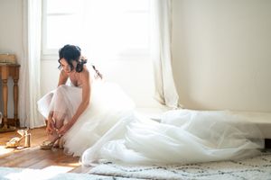 Mariée se préparant, assise près d’une fenêtre dans une chambre lumineuse, moment calme et intime avant la cérémonie.