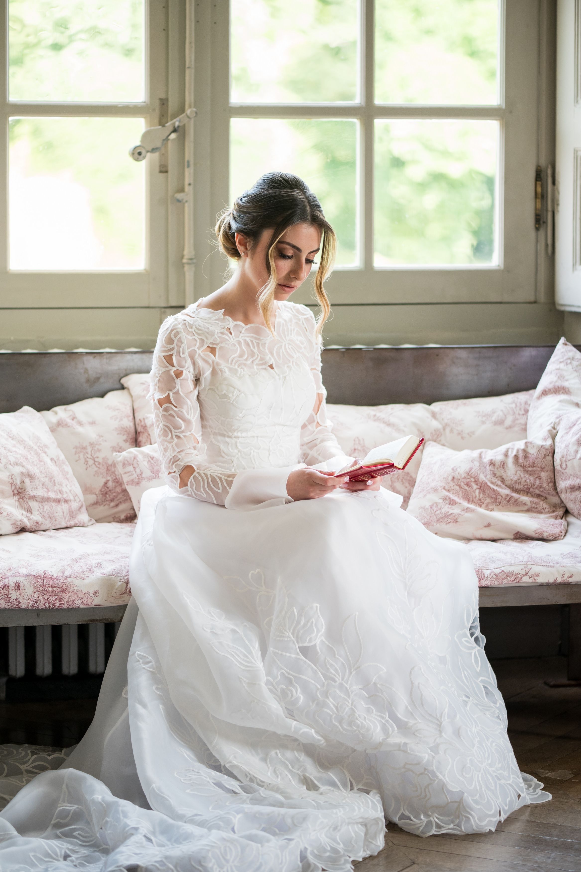 Mariée assise en robe blanche près d’une grande fenêtre, instant calme et élégant juste avant le mariage. Berakhot avant la Houppa.