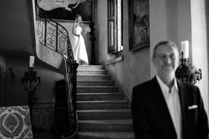 Mariée posant en haut d’un escalier en noir et blanc, composition élégante avec son père au premier plan.