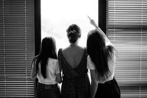 Trois femmes de dos devant une grande fenêtre en noir et blanc, moment calme et lumineux avant la cérémonie.