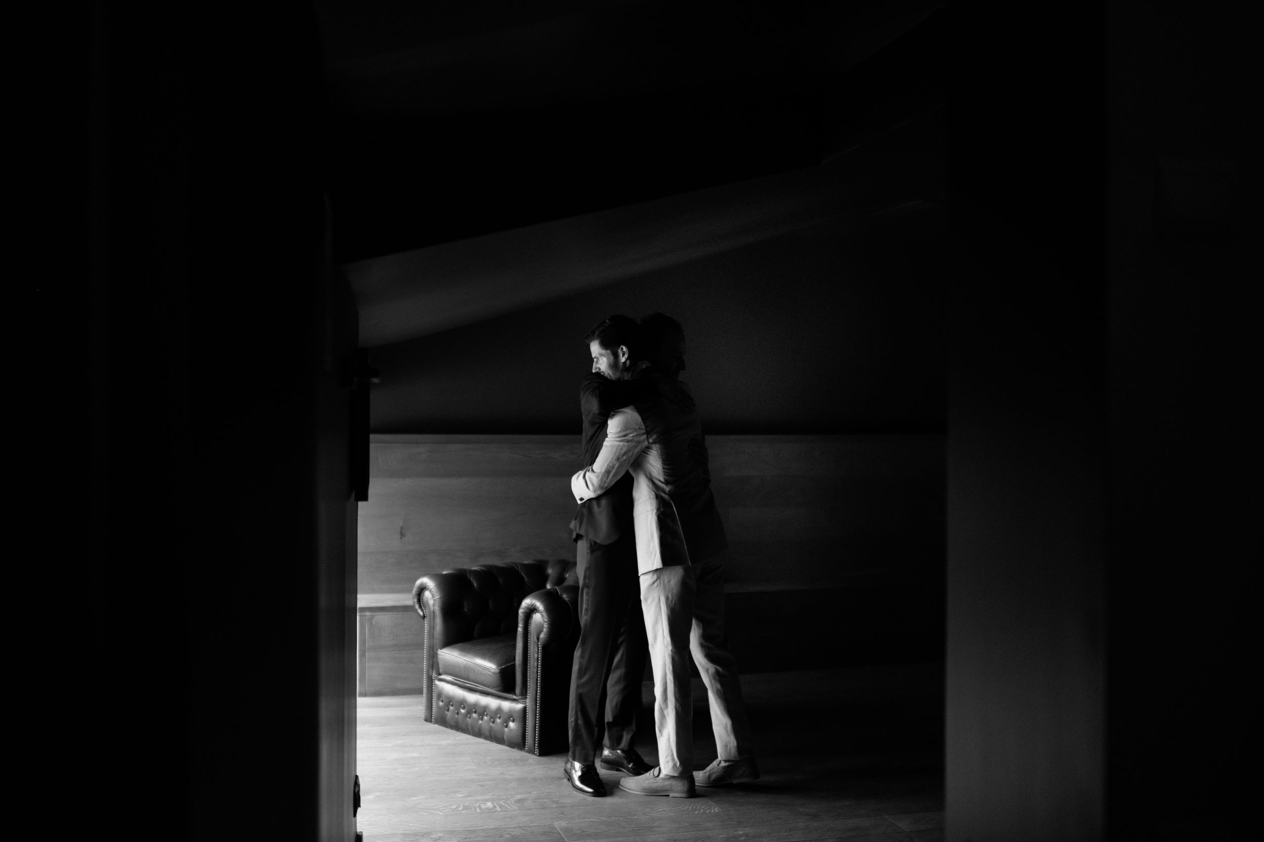 Deux amis enlacés dans une chambre sombre en noir et blanc, scène intime et cinématographique des préparatifs du Hatan, le marié.