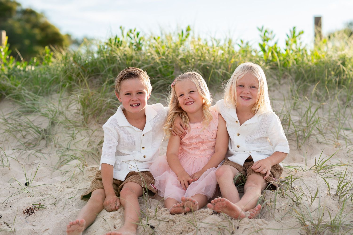 Family of five posing on driftwood log at sandy beach with grass dunes at sunset.