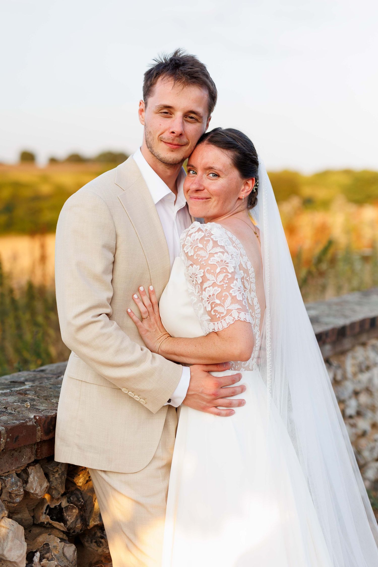 Bride and groom embracing outdoors at sunset, she in white lace gown with veil, he in beige suit, golden fields behind.