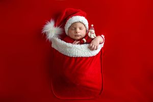 Newborn baby sleeping in a red Santa sack wearing a Santa hat, holding a tiny snowman figurine on red background.