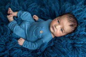 Newborn baby in blue outfit lying on fluffy blue rug, looking up with dark eyes during professional photo session.