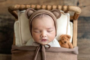 Sleeping newborn baby in bear bonnet hat lying in tiny wooden bed with teddy bear, professional newborn photography.