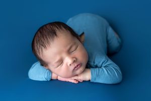 Peaceful newborn baby sleeping in froggy pose wearing blue outfit on blue background.