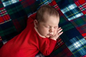 Newborn baby in red onesie sleeping peacefully on a plaid tartan blanket in a festive holiday photo.
