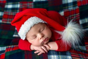 Sleeping newborn baby wearing a red Santa Claus hat and outfit on a plaid blanket.