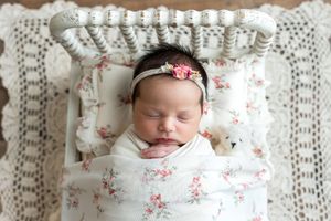 Sleeping newborn baby girl swaddled in floral wrap with pink headband in a white miniature bed.