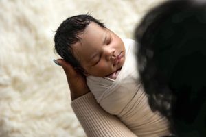 Newborn baby sleeping peacefully wrapped in a cream swaddle, cradled in loving hands on a white fluffy backdrop.