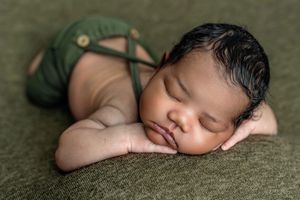 Sleeping newborn baby in green overalls posed on a green blanket in a classic froggy position.