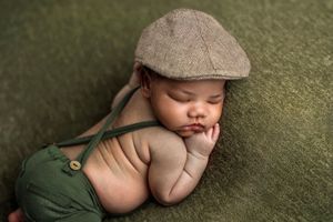 Sleeping newborn baby wearing a tan flat cap and green suspender pants on a green textured blanket.