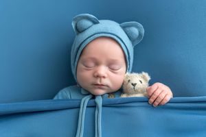 Sleeping newborn baby in blue bear hat cuddles a small teddy bear, wrapped in a soft blue blanket.