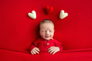 Sleeping newborn baby in red outfit surrounded by red and white felt hearts on a red background.