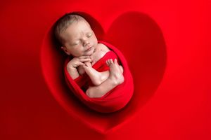 Newborn baby sleeping peacefully in a red heart-shaped nest, wrapped in red fabric on a red background.