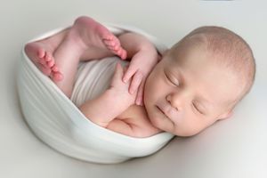 Peaceful newborn baby sleeping curled up in a white bowl wrap, showcasing tiny fingers and toes in a studio portrait.