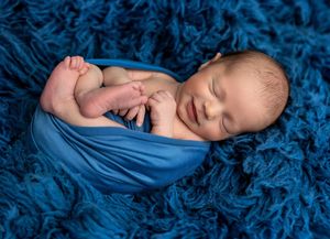 Sleeping newborn baby wrapped in blue fabric on a fluffy blue blanket in a cozy posed photo.