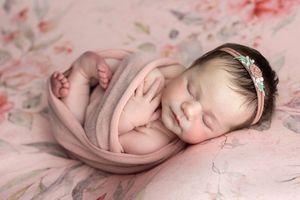 Sleeping newborn baby girl wrapped in pink swaddle with floral headband on pink floral blanket.