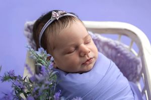 Newborn baby girl wrapped in lavender swaddle with floral headband, sleeping peacefully in white bed with purple flowers.