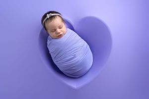 Newborn baby swaddled in purple wrap sleeping in heart-shaped cutout for studio photography.