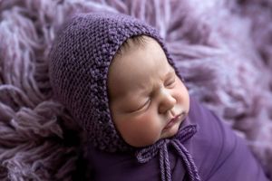 Sleeping newborn baby wrapped in purple knit bonnet and swaddle on a soft lavender fur blanket.
