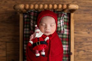 Sleeping newborn in red knit wrap and hat holding a Santa Claus toy in a small wooden bed with plaid bedding.