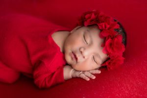 Newborn baby girl sleeping peacefully in red outfit and floral headband on red background.