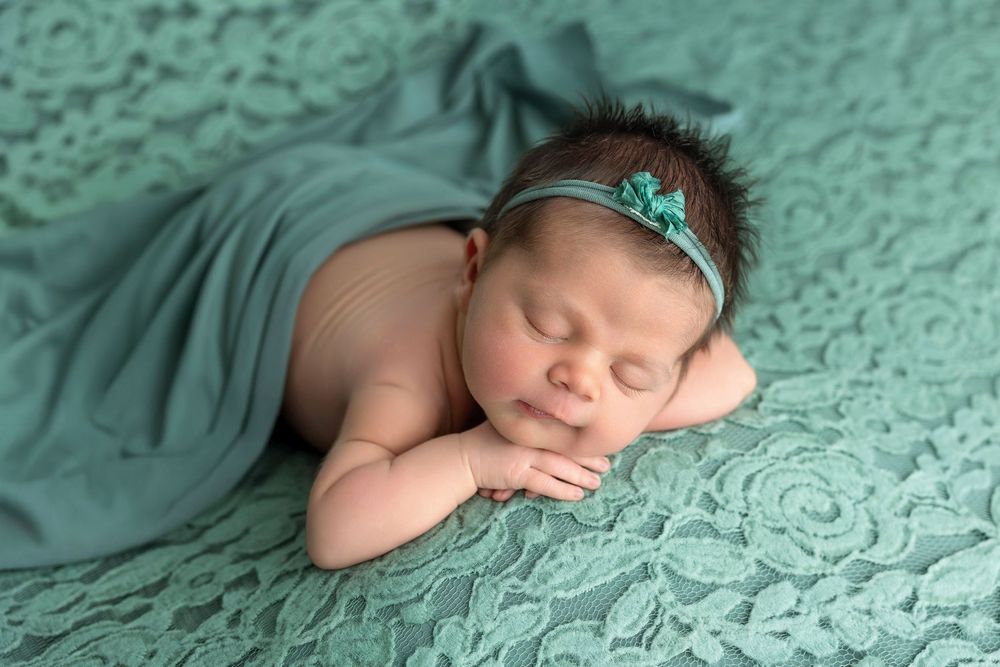 Peaceful newborn baby sleeping on teal lace blanket, wearing matching floral headband, wrapped in soft turquoise swaddle.
