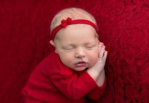 Sleeping newborn baby in red outfit and bow headband on red lace background.