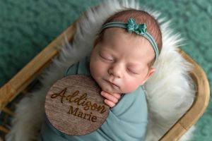 Newborn baby girl named Addison Marie sleeping in basket with teal wrap and floral headband beside engraved wood name disc.