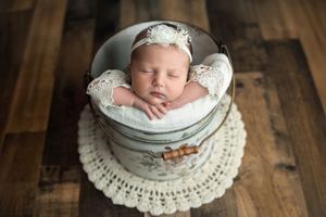 Sleeping newborn baby girl in lace outfit and floral headband posed in vintage metal bucket on wood floor.