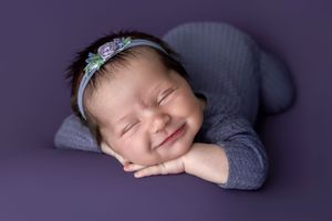 Smiling newborn baby girl in gray wrap and floral headband, sleeping peacefully on purple background.