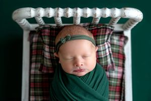 Newborn baby swaddled in green wrap with floral headband, sleeping peacefully in a white mini bed with plaid bedding.