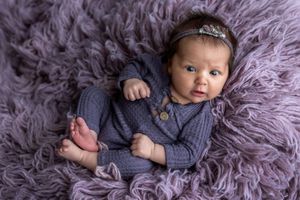 Newborn baby girl in purple knit romper with floral headband lying on fluffy lavender fur backdrop.
