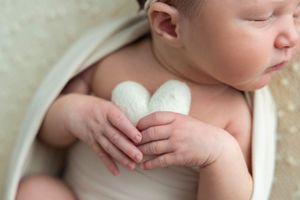 Sleeping newborn baby holding a small white heart-shaped prop, wrapped in a cream swaddle blanket.