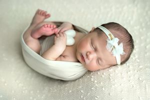 Sleeping newborn baby girl in white bowl with angel wings and bow headband on white textured blanket.
