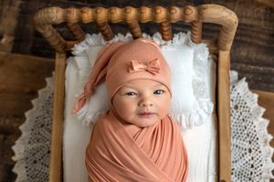 Newborn baby swaddled in peach wrap wearing matching bow hat, lying in a wooden mini bed with white lace.